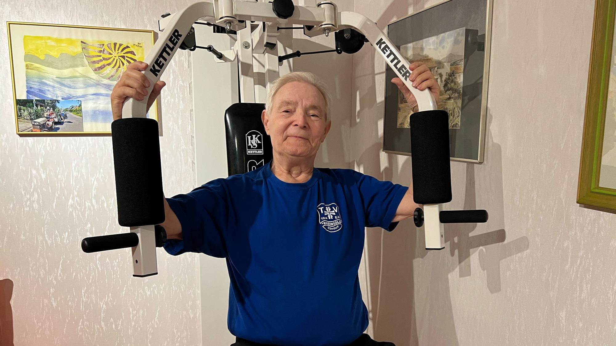 Ein älterer Mann mit grauem Haar sitzt auf einem Fitnessgerät, das die Armmuskulatur stärkt.