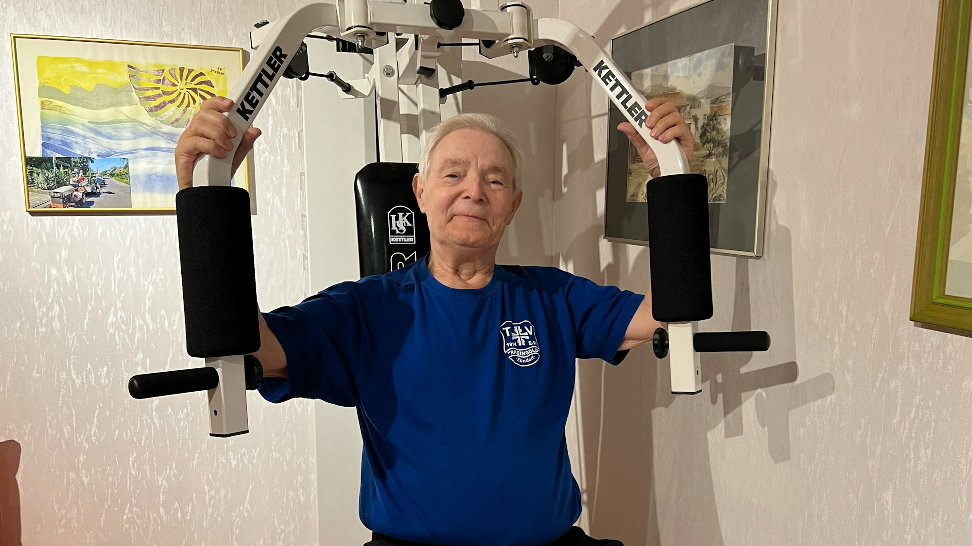 Ein älterer Mann mit grauem Haar sitzt auf einem Fitnessgerät, das die Armmuskulatur stärkt.