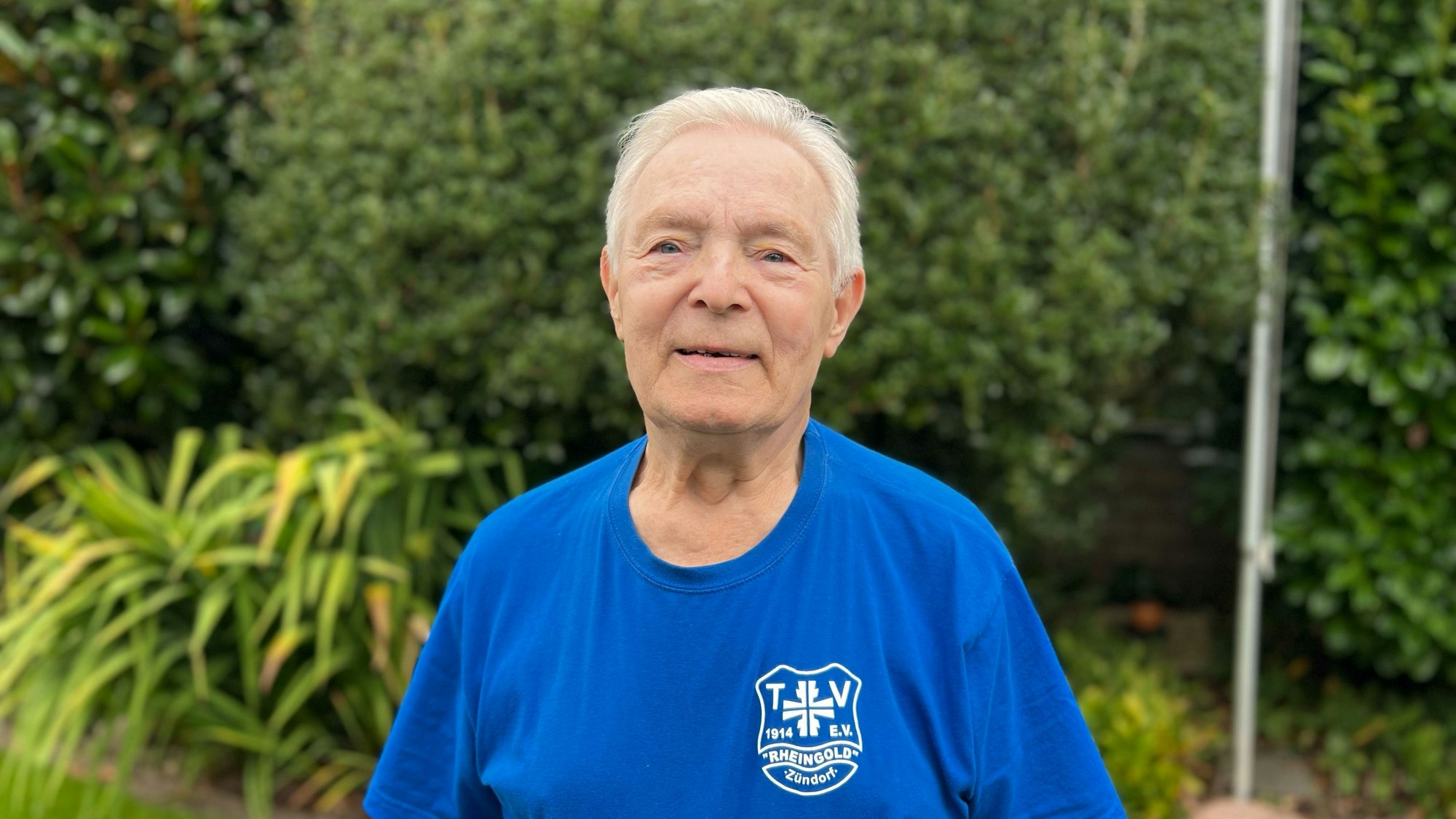 Ein älterer Mann im blauen T-Shirt steht in einem Garten.