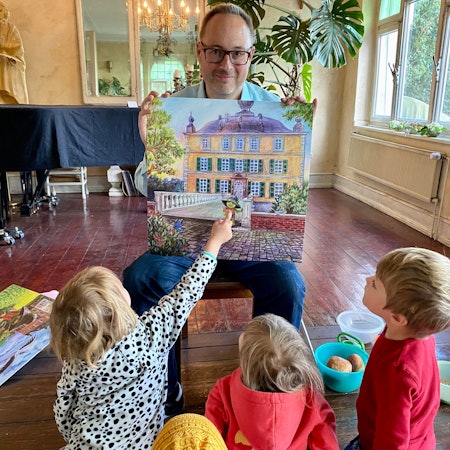 Der Autor hält eine Zeichnung in der Hand, auf der grüne Kobold vor einem prächtigen Gebäude zu sehen ist.