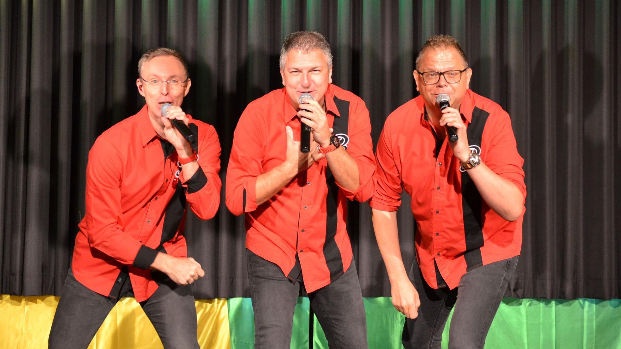Die drei Sänger der Band „Bremsklötz“ tragen identische rote Hemden und singen in Mikrofone.