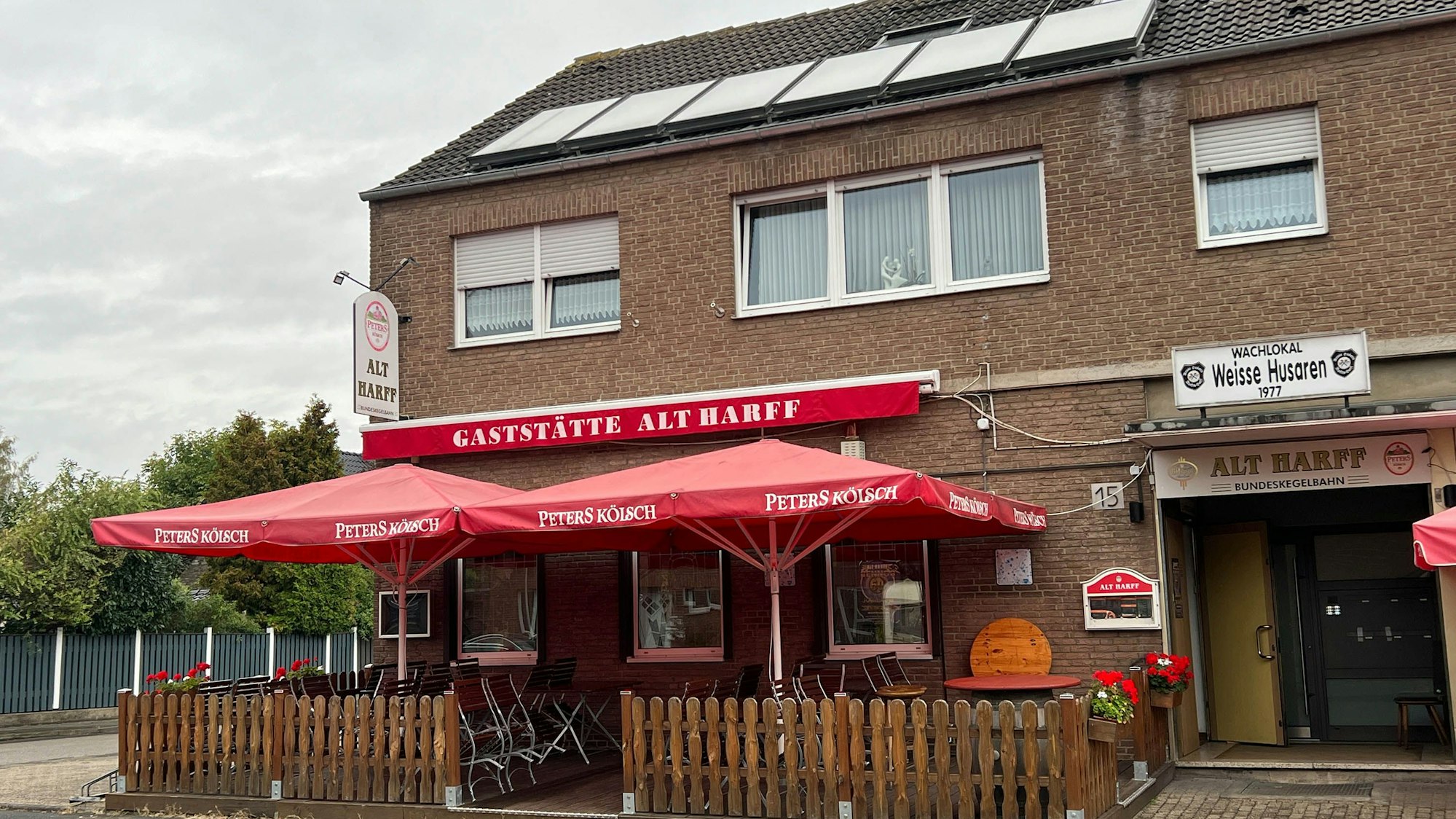 Vor einer Gaststätte stehen eingerahmt von einem Jägerzaun Tische, Stühle und rote Sonnenschirme.