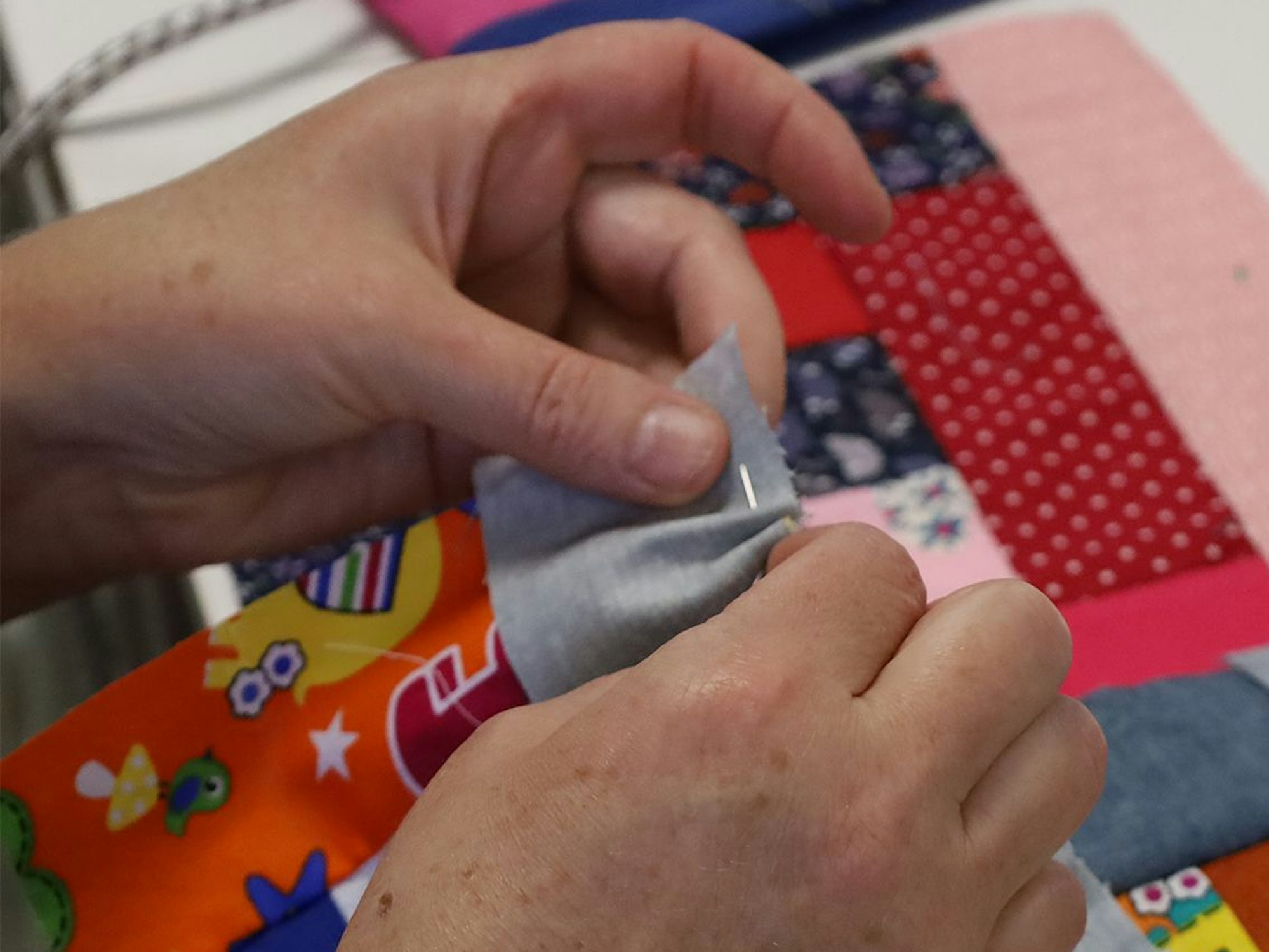 Das Bild zeigt die Hände einer Frau, die mit Nadel und Faden bunte Stoff-Rechtecke zusammennäht.