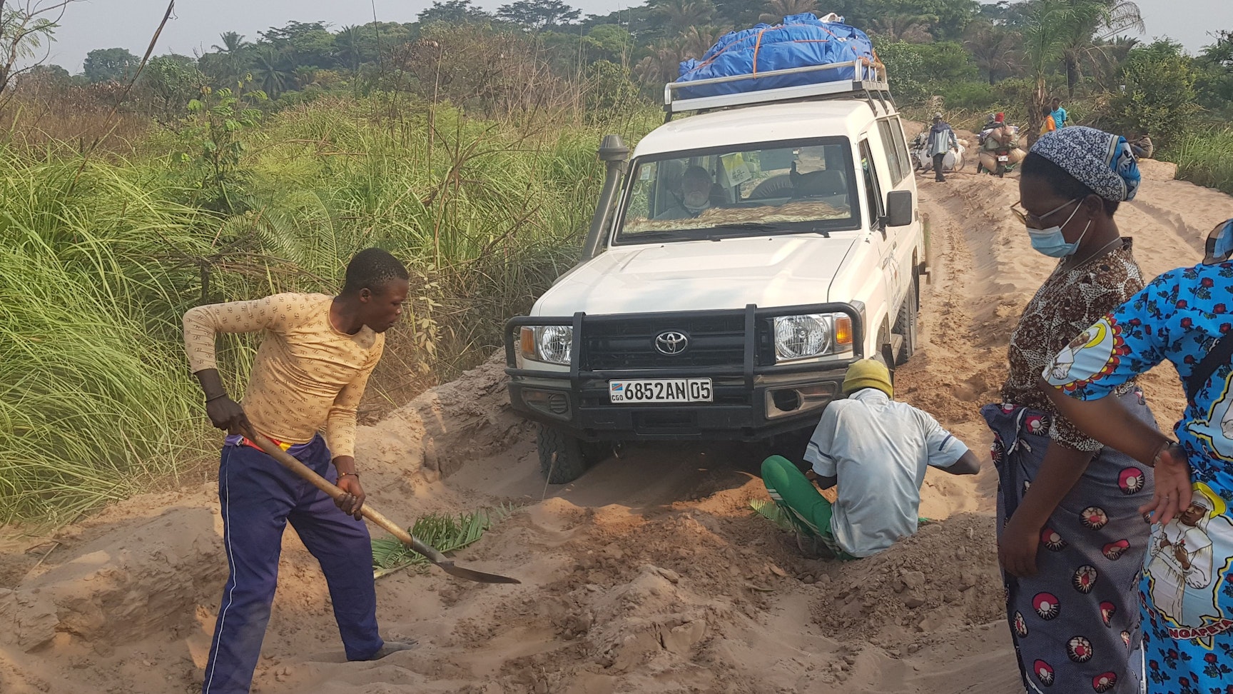 Der Wagen, mit dem Hans Jagsch und Manfred Janz durch den Kongo fuhren, steckt im Sand fest. Zwei Männer, einer mit Schaufel, graben das Fahrzeug frei.