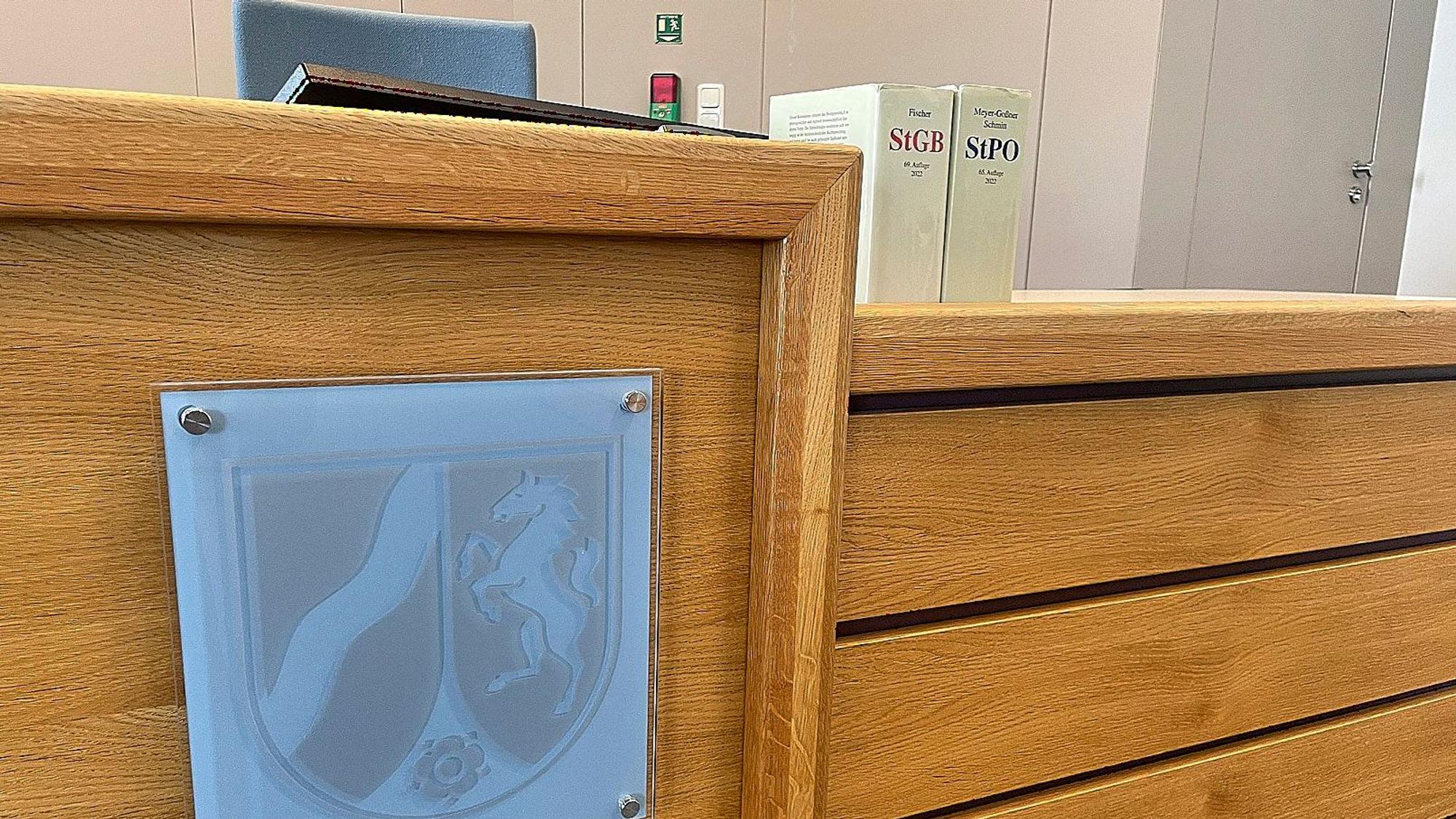 Das Foto zeigt den Richtertisch im Amtsgericht in Gemünd. In Glas ist das Wappen des Bundeslandes Nordrhein-Westfalen angebracht, auf dem Tisch stehen zwei Bücher mit Gesetzestexten.