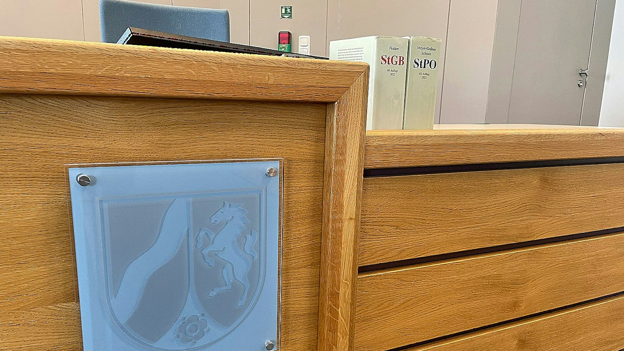 Das Foto zeigt den Richtertisch im Amtsgericht in Gemünd. In Glas ist das Wappen des Bundeslandes Nordrhein-Westfalen angebracht, auf dem Tisch stehen zwei Bücher mit Gesetzestexten.