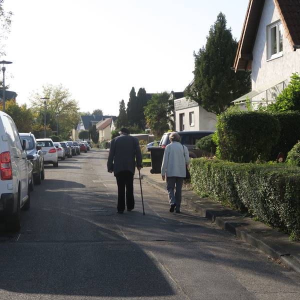 Ein älteres Paar geht auf der Fahrbahn einer Straße. Der Gehweg ist sehr schmal.