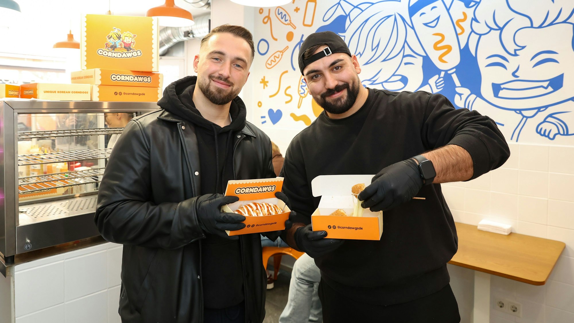 Die zwei Männer halten Packungen mit den Corndogs hoch