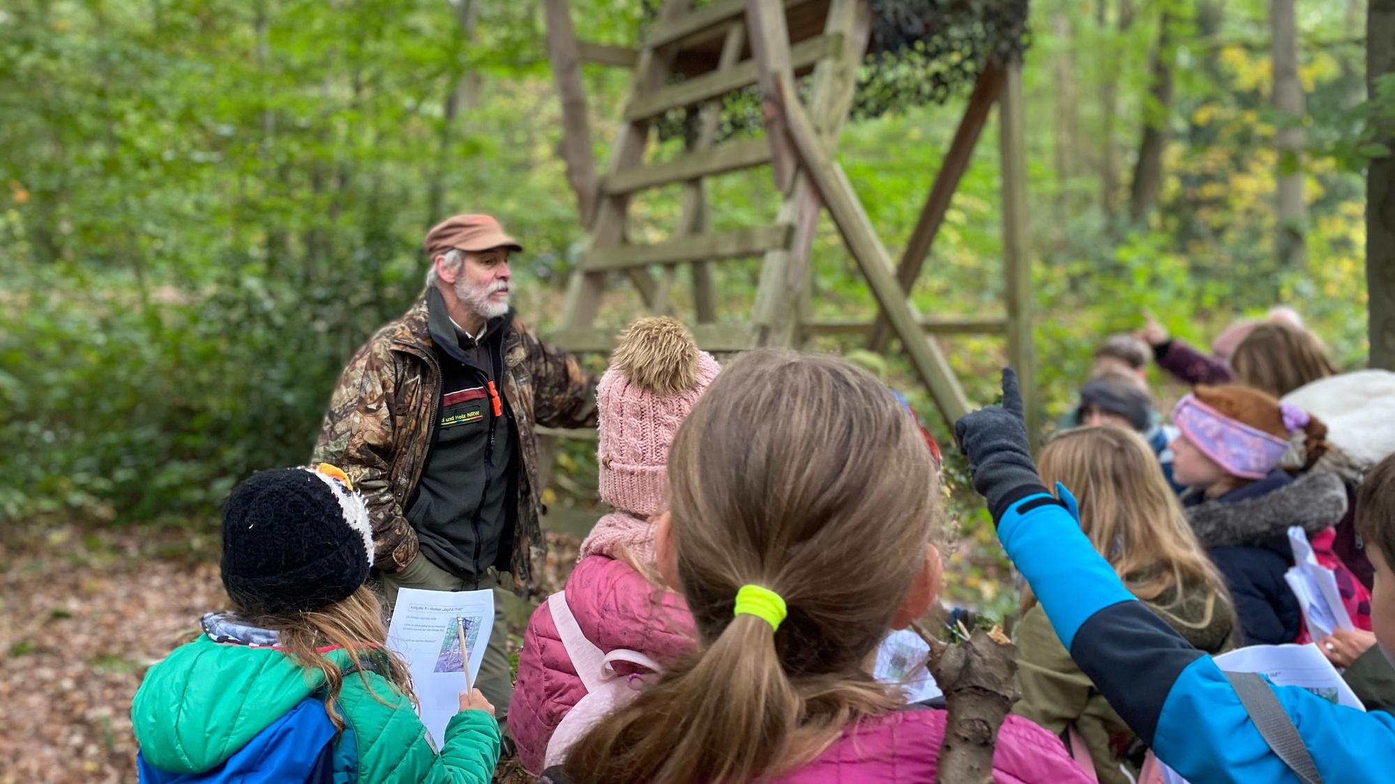 Grundschulkinder stehen in einer Traube im Wald, schauen Euskirchener Förster Heinz Benden an und heben ihre Finger, um eine Frage zu stellen.
