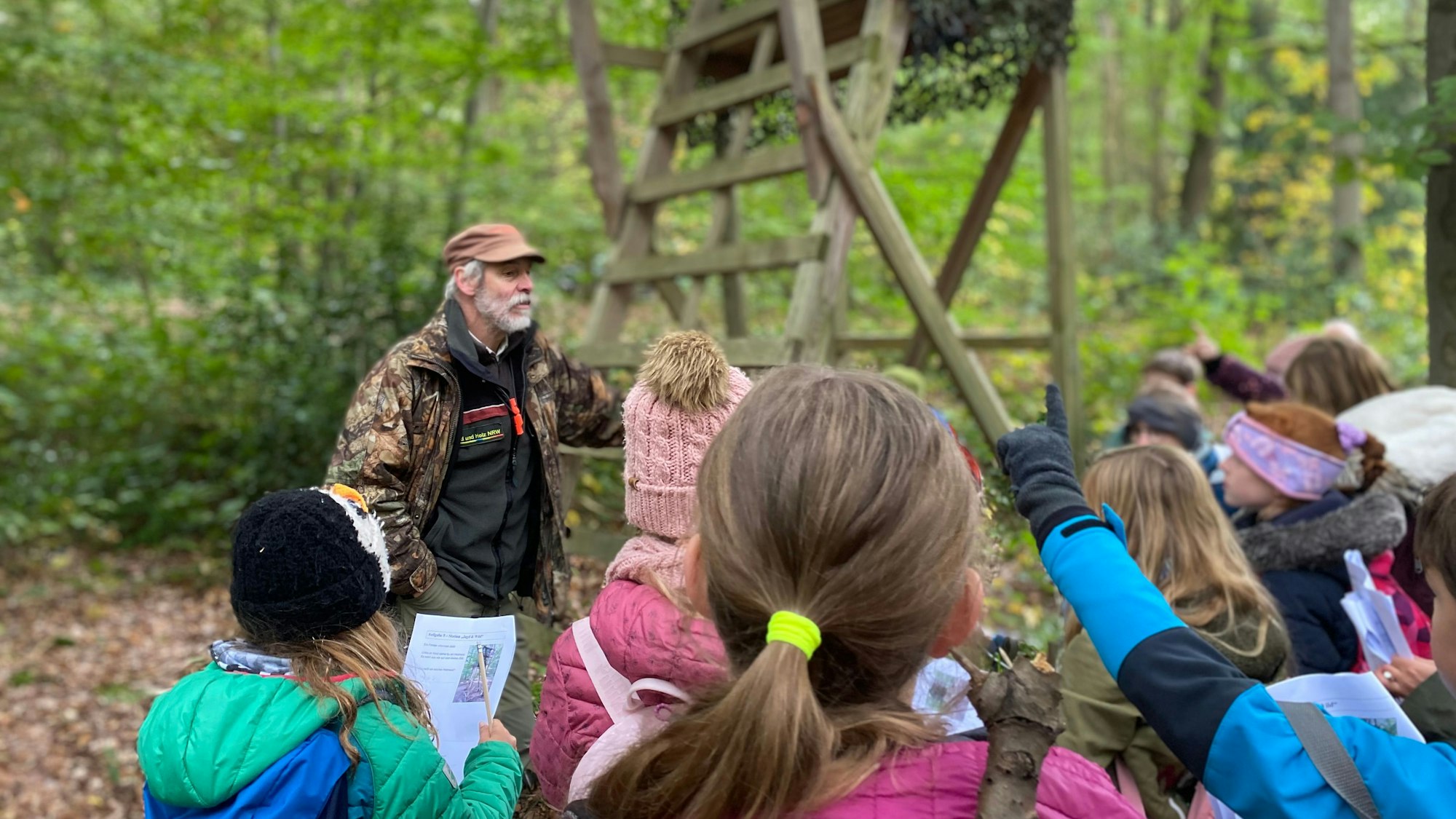 Grundschulkinder stehen in einer Traube im Wald, schauen Euskirchener Förster Heinz Benden an und heben ihre Finger, um eine Frage zu stellen.