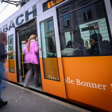 Eine Frau in einer rosa Jacke steigt an einer Bahnhaltestelle in eine Bahn der SWB ein. Weitere Personen am Bahngleis sind nicht erkennbar. Ein Mann schaut rechts im Bild aus dem Bahnfenster.
