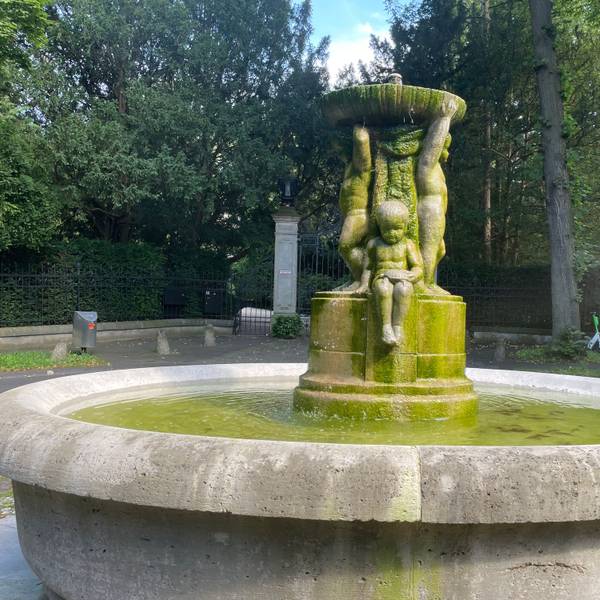 Ein steinerner Brunnen mit kindlichen Figuren steht in einer Parkanlage.