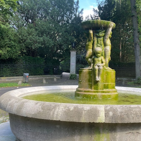 Ein steinerner Brunnen mit kindlichen Figuren steht in einer Parkanlage.