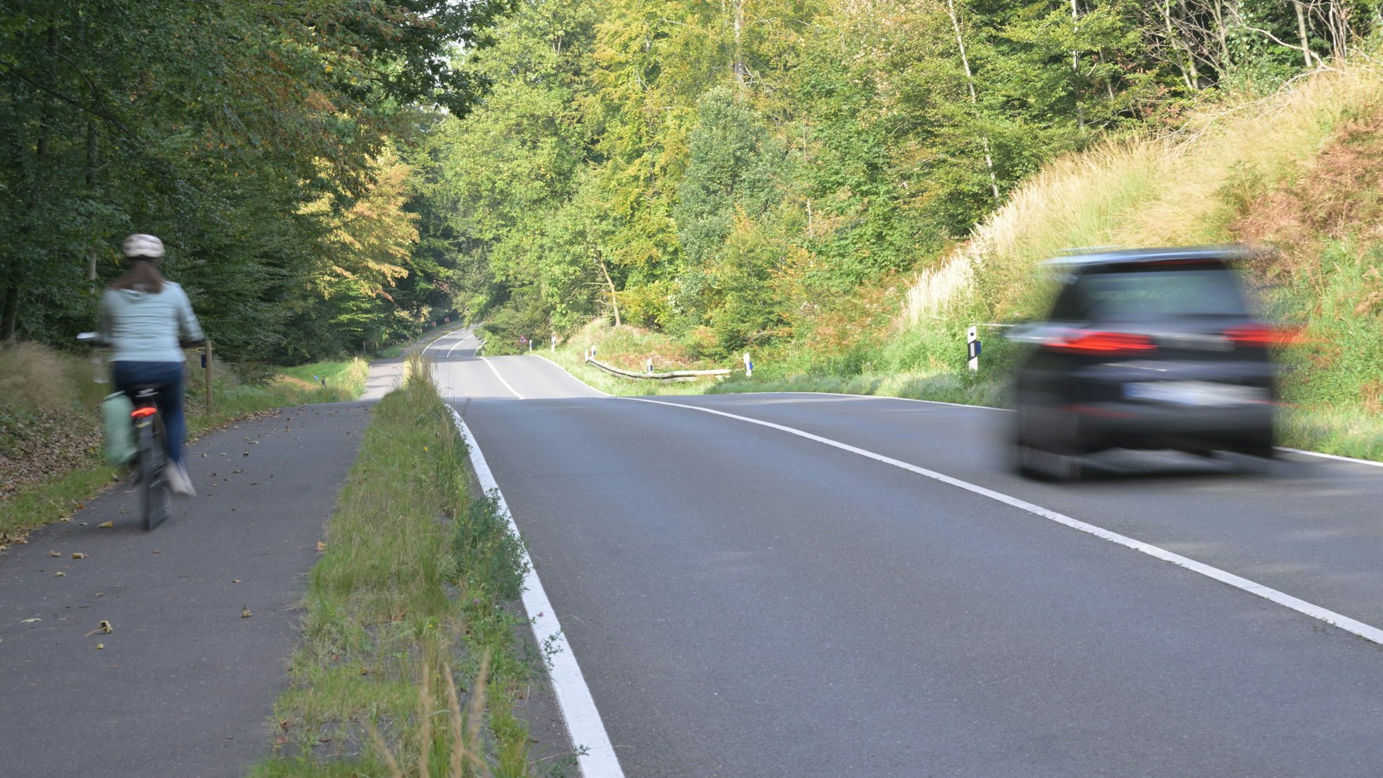 Ein Auto fährt über eine Landstraße, daneben fährt ein Fahrrad auf einem Radweg.