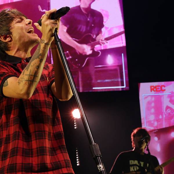 Tomlinson trägt ein rot-schwarz-kariertes T-Shirt. Er singt in ein Mikrofon. Im Hintergrund Bildschirme, die ebenfalls den Auftritt zeigen, aber pink verfärbt sind.