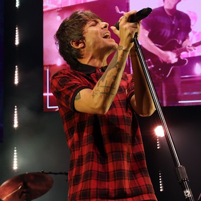 Tomlinson trägt ein rot-schwarz-kariertes T-Shirt. Er singt in ein Mikrofon. Im Hintergrund Bildschirme, die ebenfalls den Auftritt zeigen, aber pink verfärbt sind.