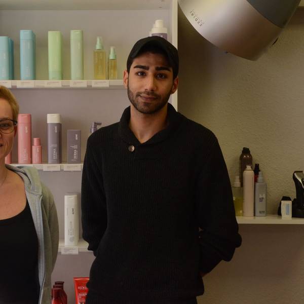 Ein Frau mit kurzen blonden Haaren und ein dunkelhaariger Mann in schwarzer Kleidung und mit einer schwatrzen Baseball-Kappe stehen vor einem Regal mit Friseurprodukten.