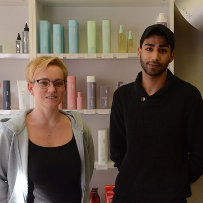 Ein Frau mit kurzen blonden Haaren und ein dunkelhaariger Mann in schwarzer Kleidung und mit einer schwatrzen Baseball-Kappe stehen vor einem Regal mit Friseurprodukten.