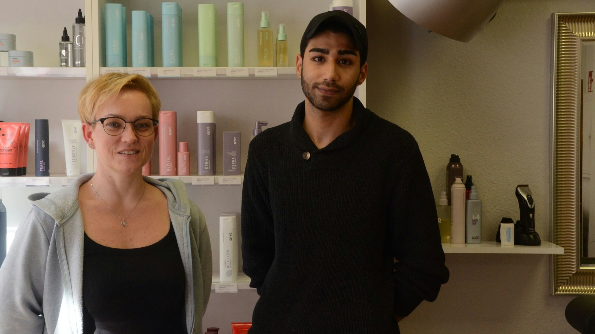 Ein Frau mit kurzen blonden Haaren und ein dunkelhaariger Mann in schwarzer Kleidung und mit einer schwatrzen Baseball-Kappe stehen vor einem Regal mit Friseurprodukten.