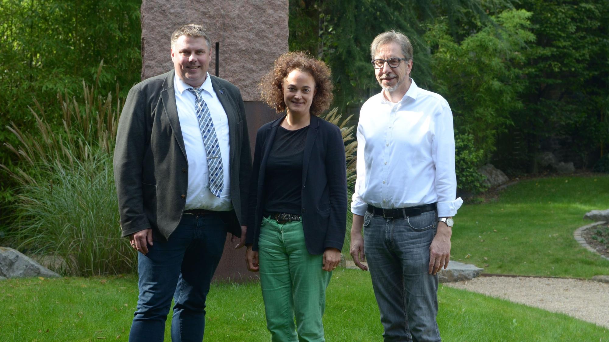 Michael Mombaur (l.), Barbara Luke und Dr. Hubertus Rüber stehen im Freien vor einer Steinskulptur.