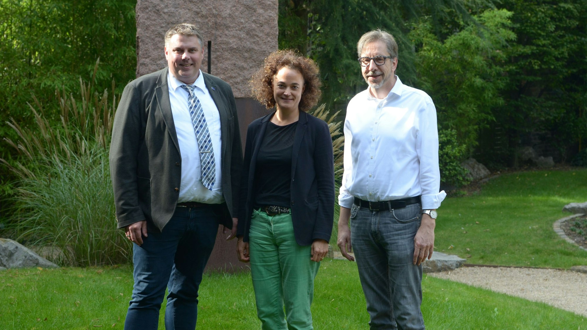 Michael Mombaur (l.), Barbara Luke und Dr. Hubertus Rüber stehen im Freien vor einer Steinskulptur.