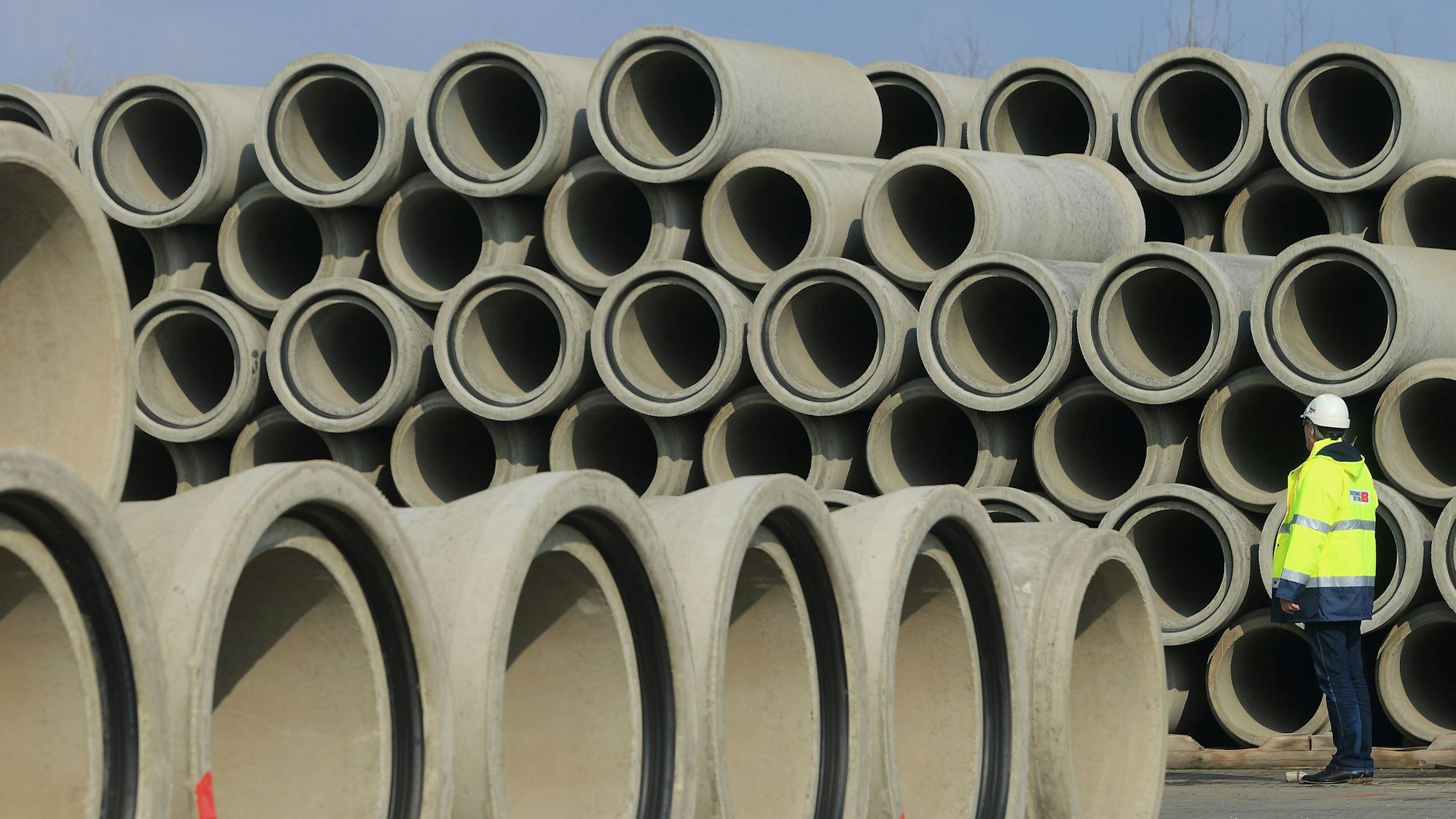 Zu einer hohen Wand aufgestapelte Kanalrohre. Davor steht ein Bauarbeiter mit einer gelb reflektierenden Jacke und einem Helm auf dem Kopf.