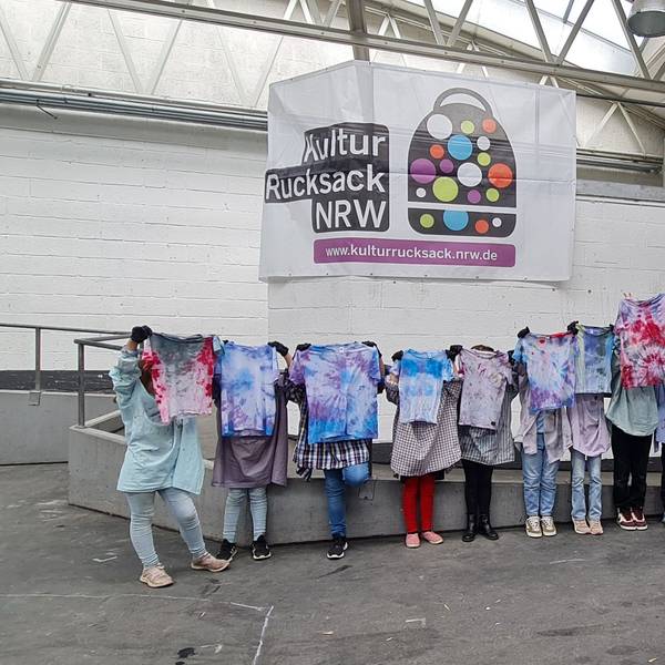 Jugendliche halten Batik-Shirts hoch, ihre Köpfe sind dahinter nicht zu sehen.