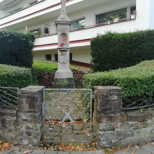 Ein steinernes Wegekreuz in einer Grünanlage vor einem Mehrfamilienhaus.