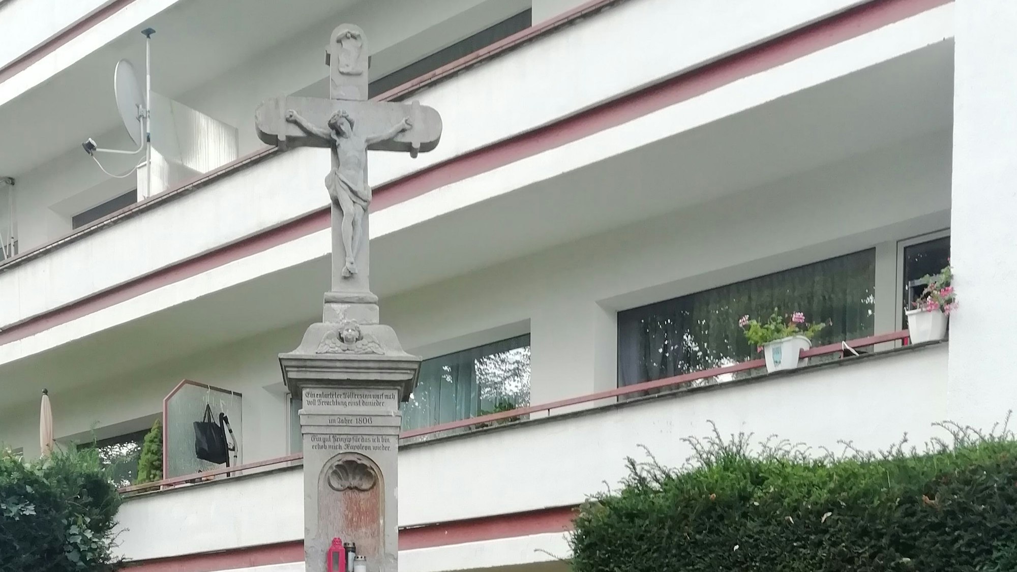 Ein steinernes Wegekreuz in einer Grünanlage vor einem Mehrfamilienhaus.