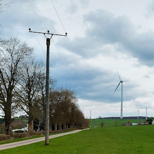 Auf einer Wiese sollen Photovoltaik-Module aufgebaut werden. Im Hintergrund sind ein Bauernhof und ein Windrad zu sehen, links eine Stromleitung.
