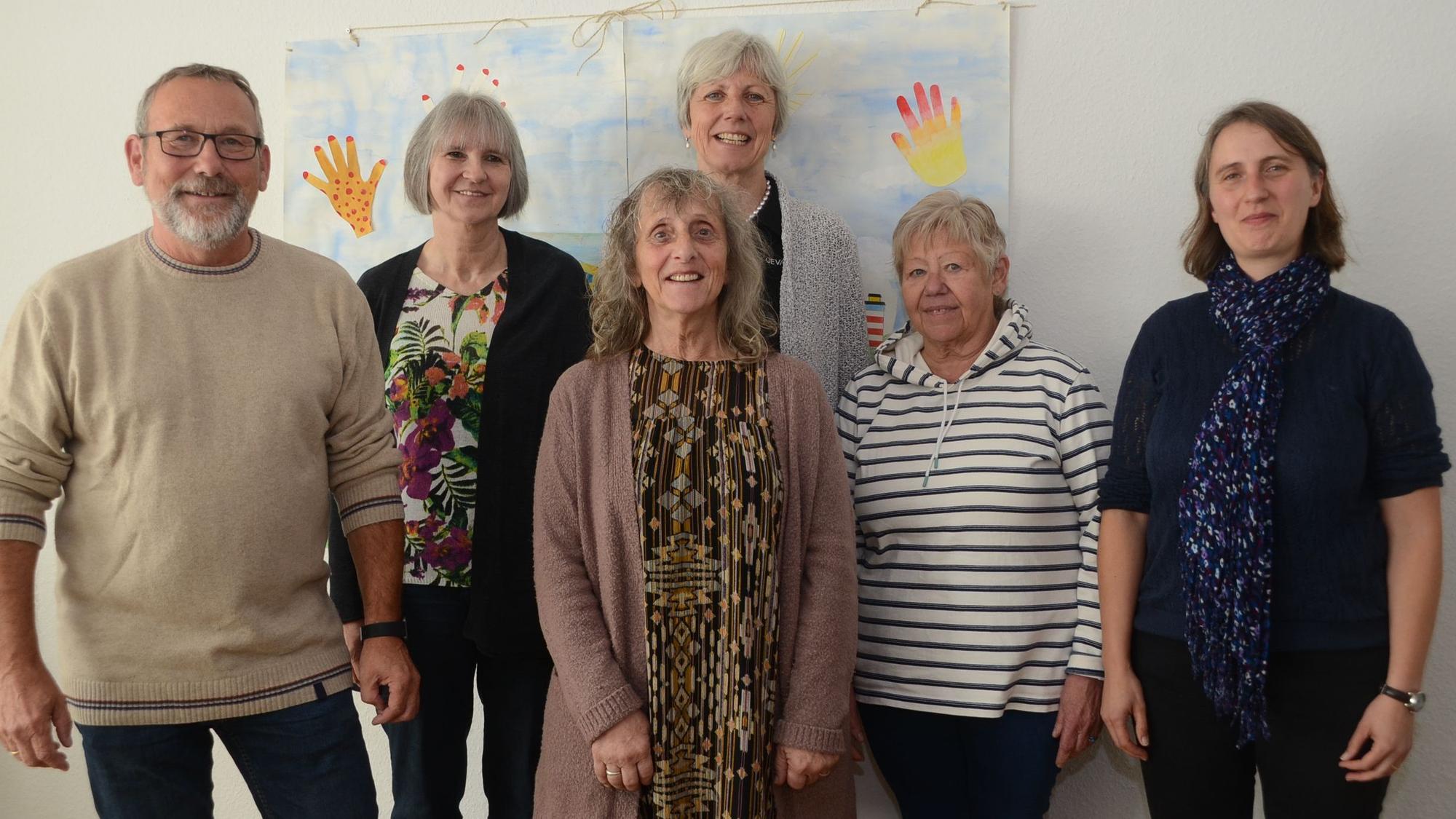 Ein Mann und fünf Frauen stehen vor einem gemalten Bild an einer weiß gestrichenen Wand.