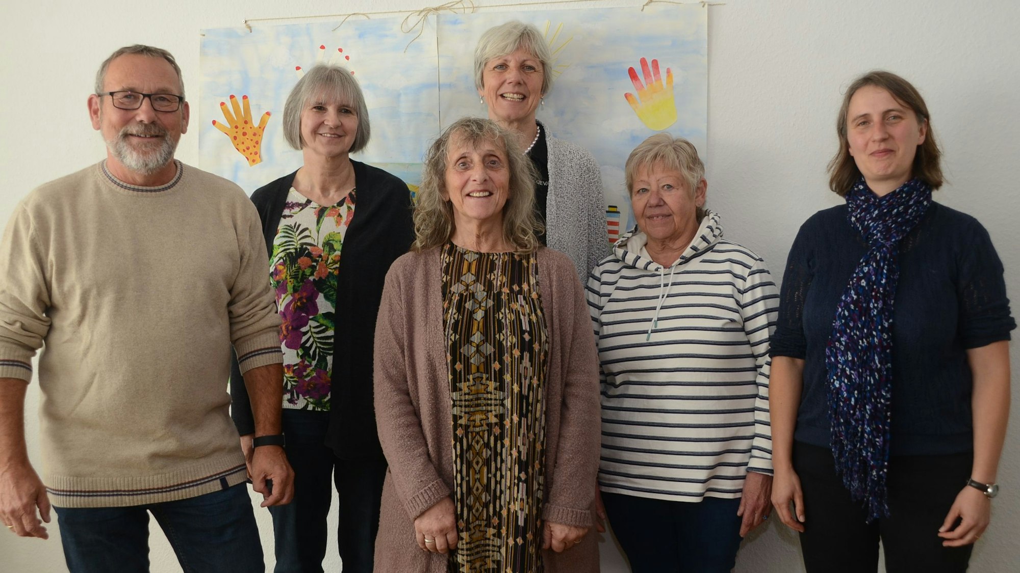 Ein Mann und fünf Frauen stehen vor einem gemalten Bild an einer weiß gestrichenen Wand.