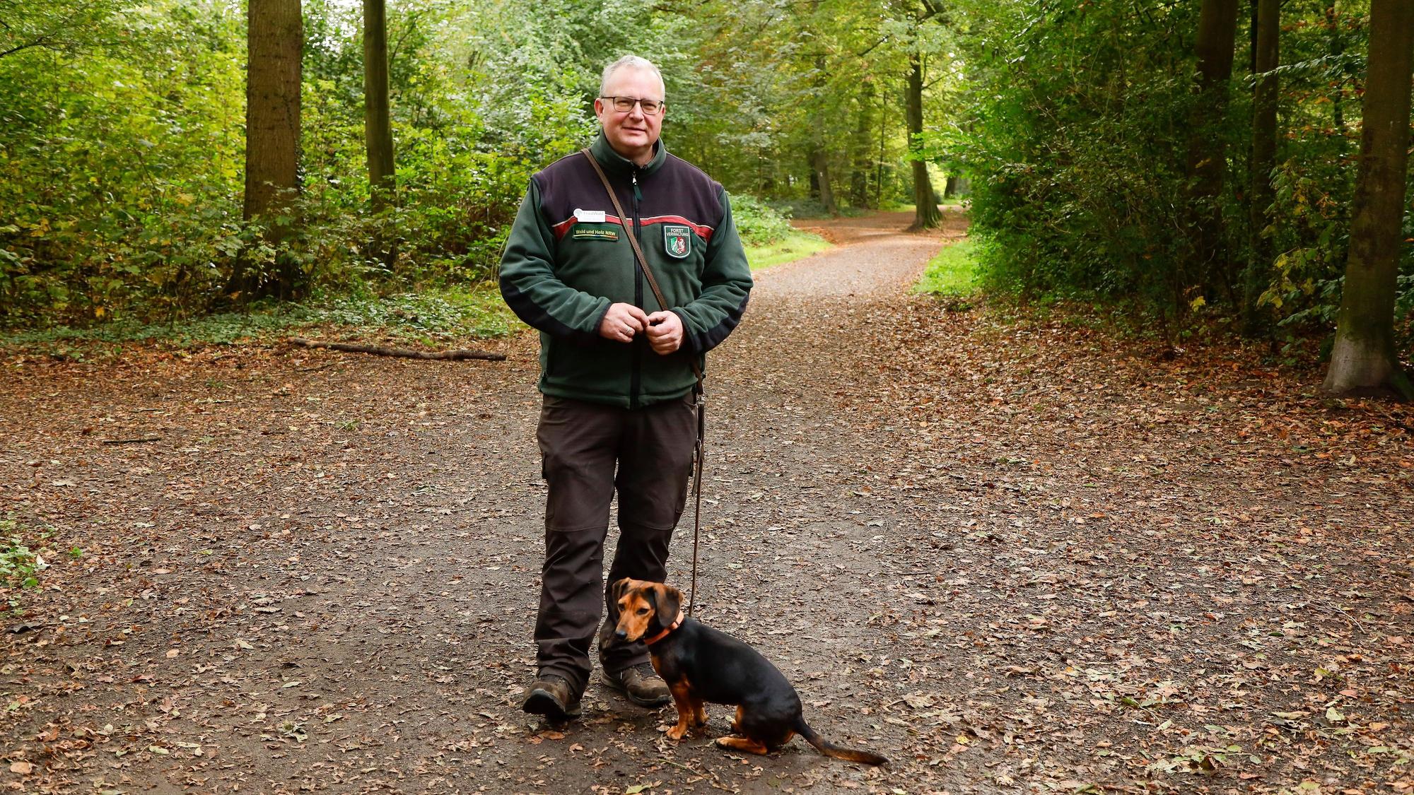 Der Förster steht in grüner Fleecejacke des Landesbetriebs auf einem Waldweg, die kleine Hündin an der Leine sitzt vor ihm.