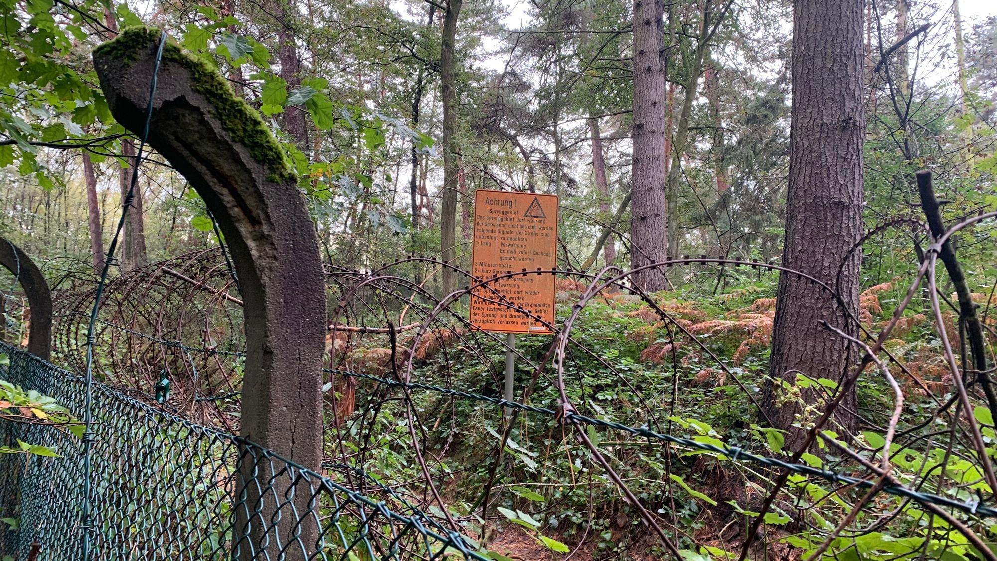 Ein alter Zaun mit Stacheldraht in einem Waldstück; im Hintergrund ein orange Warnschild.