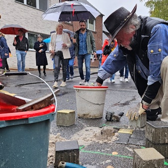 Ein Mann mit Hut kniet auf dem Gehweg und verlegt Pflasterstein, Menschen mit Regenschirmen beobachten ihn dabei.