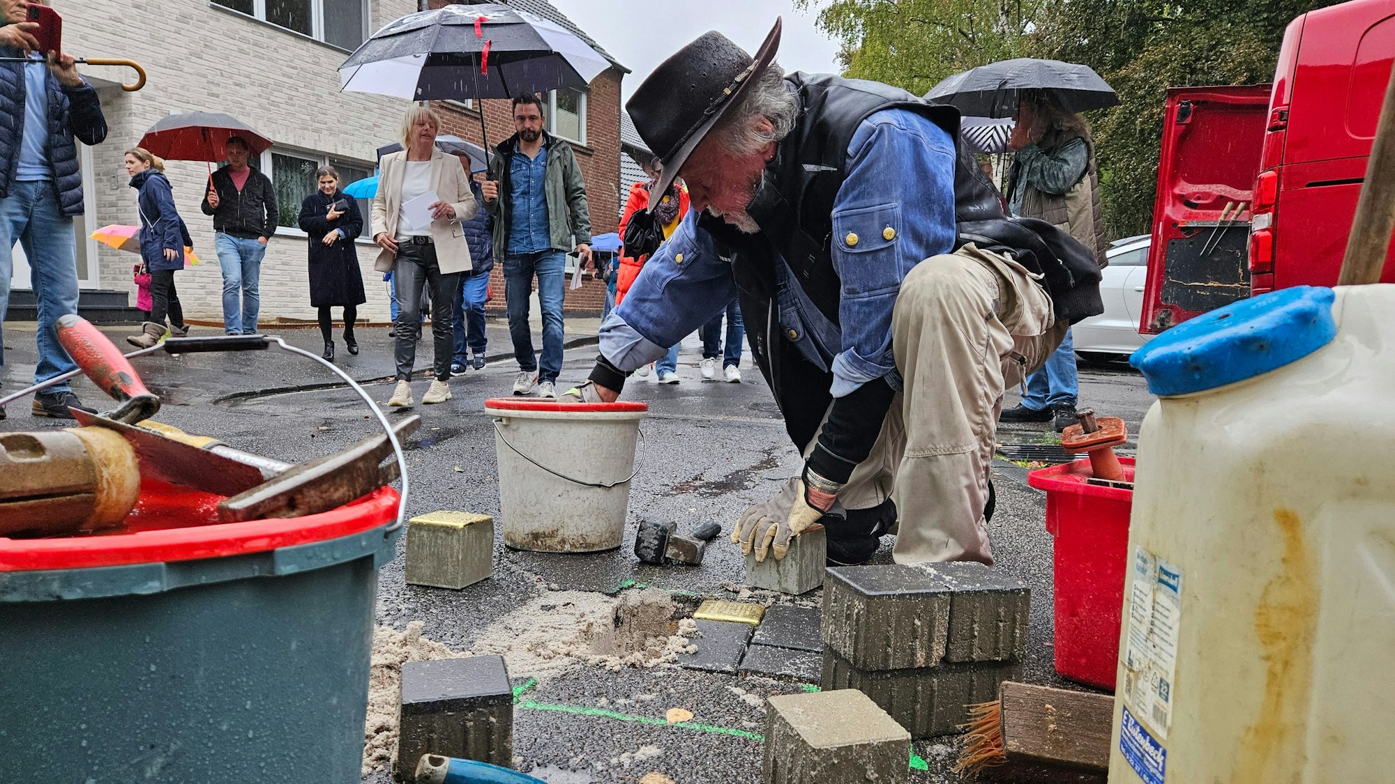 Ein Mann mit Hut kniet auf dem Gehweg und verlegt Pflasterstein, Menschen mit Regenschirmen beobachten ihn dabei.
