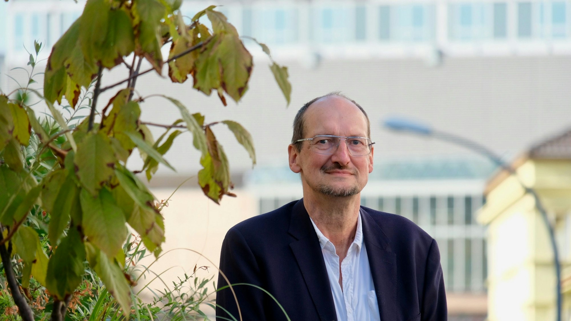Ein Mann ist im Halbporträt zu sehen, im Hintergrund ist das Gebäude der Kölner Uniklinik sichtbar.
