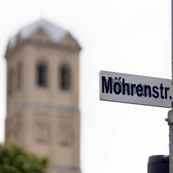An einem Straßenmast sind zwei Straßenschilder, „Mohrenstraße“ und „Gereonstraße“, angebracht, wobei erstere von Unbekannten in „Möhrenstraße“ umgewandelt wurde. Im Bildhintergrund ist die Kirche Sankt Gereon zu sehen.