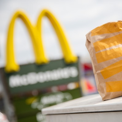 Eine McDonald’s To-Go-Tüte und ein To-Go-Becher stehen vor einer McDonald’s-Filiale.