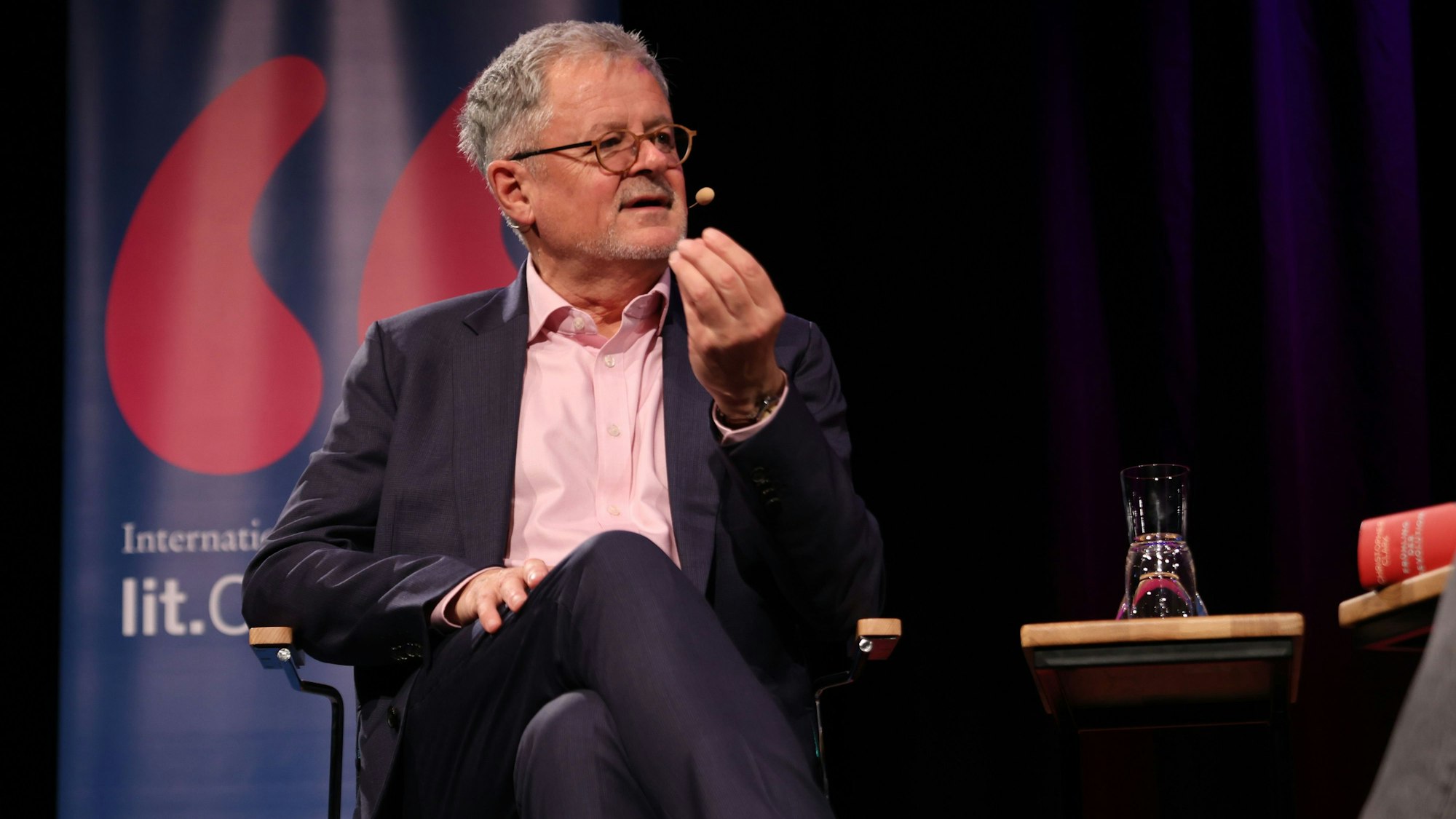 Christopher Clark trägt einen dunklen Anzug und ein rosa Hemd. Er gestikuliert mit der Hand und hat die Beine überschlagen. Hinter ihm das Banner der lit.Cologne.