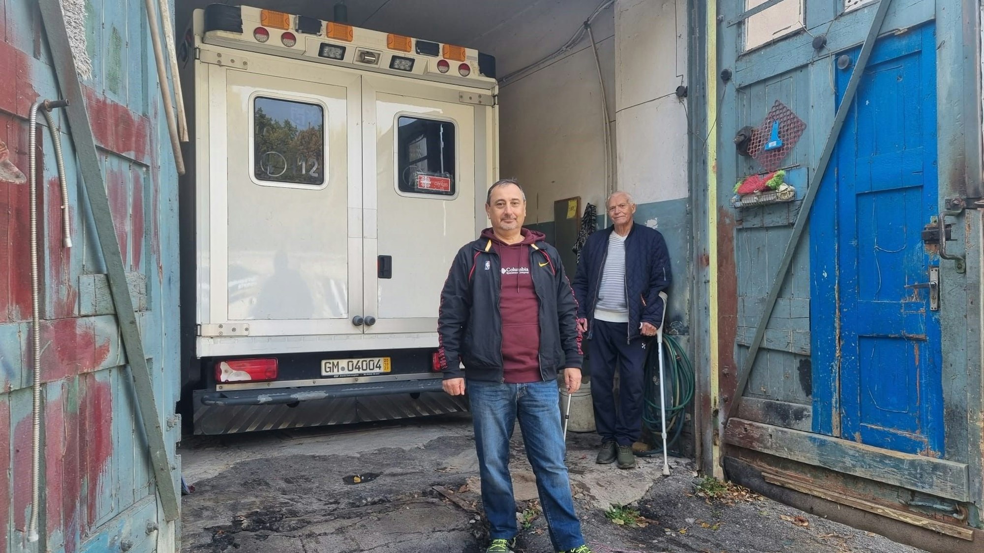 Ein Rettungswagen steht in einer Garage. Zwei Männer, einer mit Krücken, stehen davor.