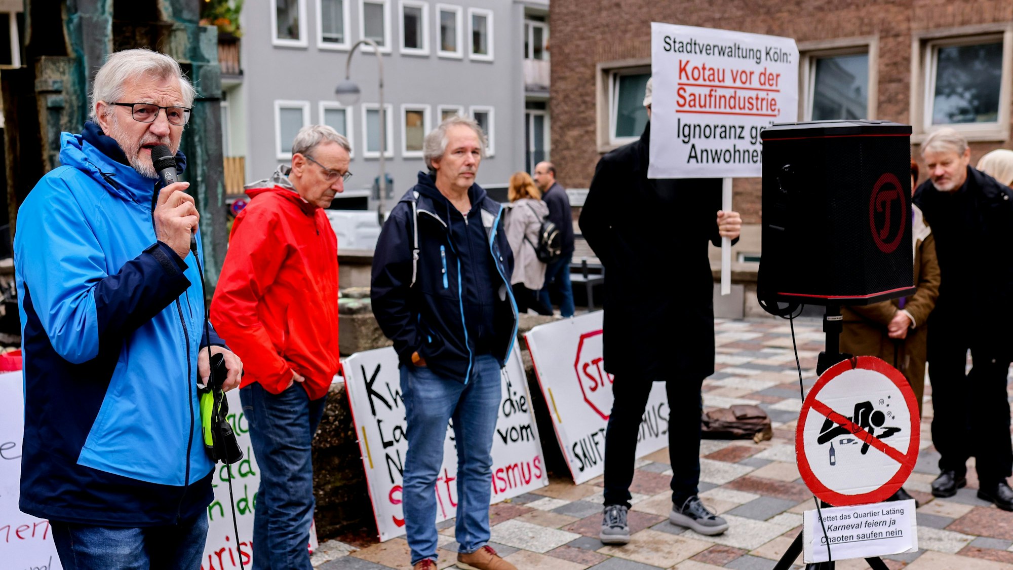 Mehrere Menschen mit Protestschildern, ein Mann spricht in ein Mikrofon.