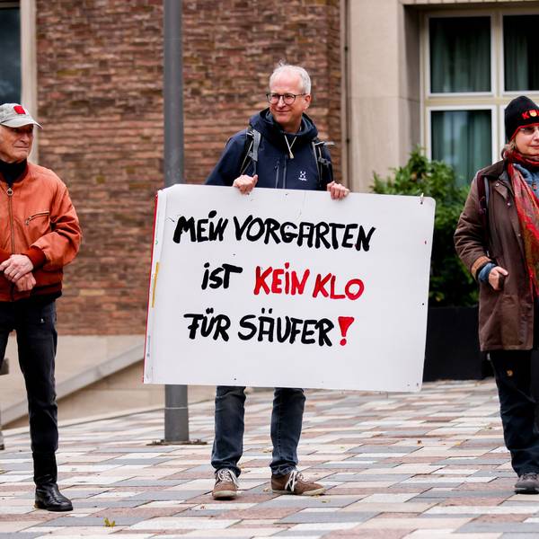 Zwei Männer und eine Frau stehen nebeneinander, ein Mann hat ein Protestschild in den Händen.