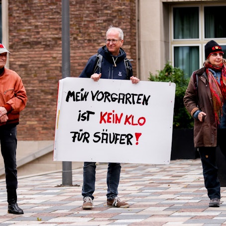 Zwei Männer und eine Frau stehen nebeneinander, ein Mann hat ein Protestschild in den Händen.