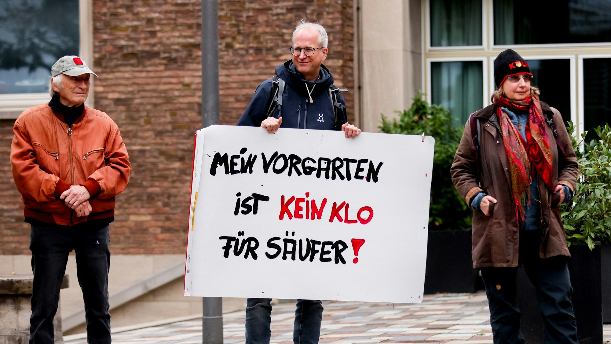 Zwei Männer und eine Frau stehen nebeneinander, ein Mann hat ein Protestschild in den Händen.