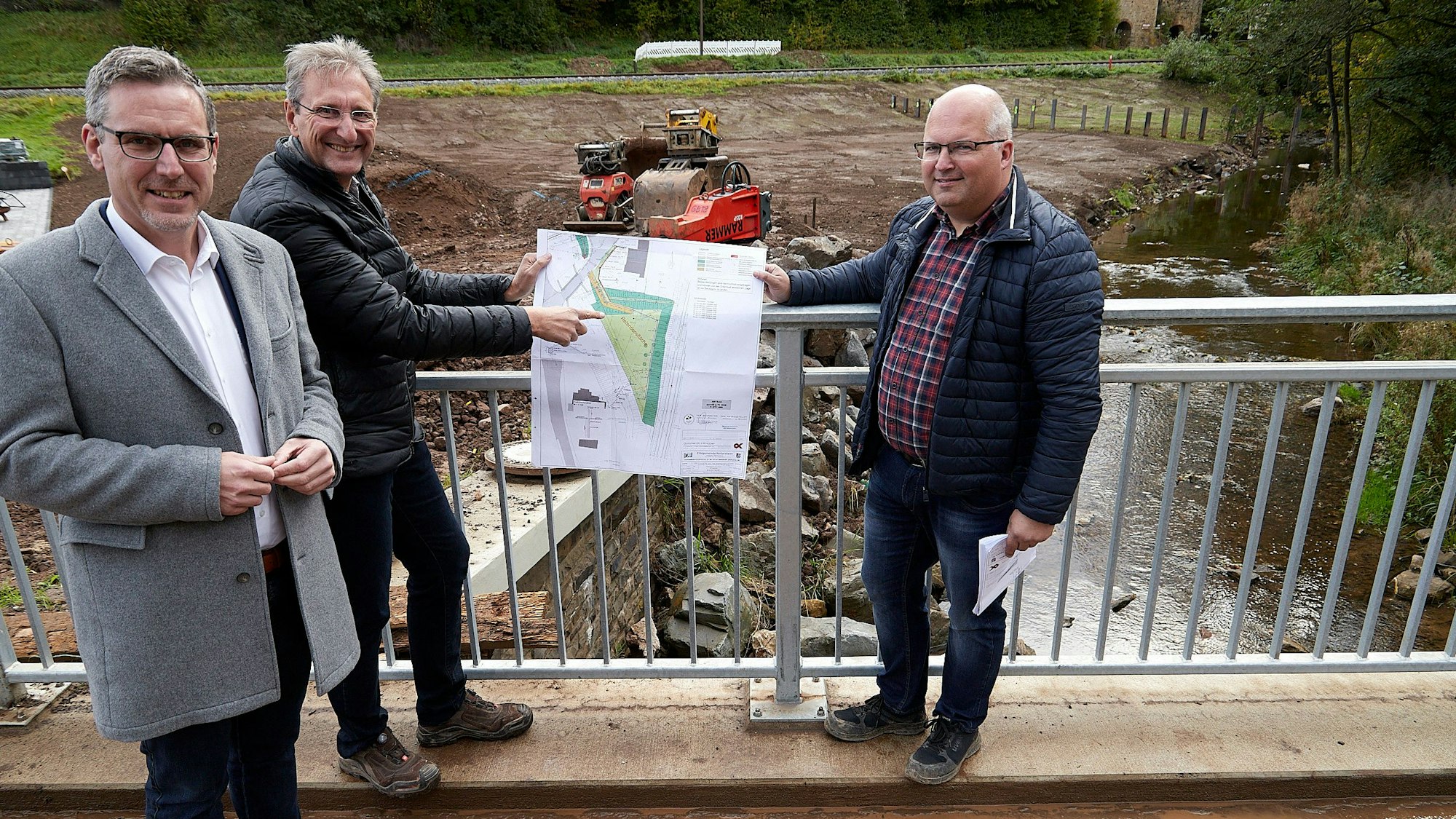 Mit einem Plan zeigen die drei auf einer Brücke, welche Maßnahmen getroffen werden.