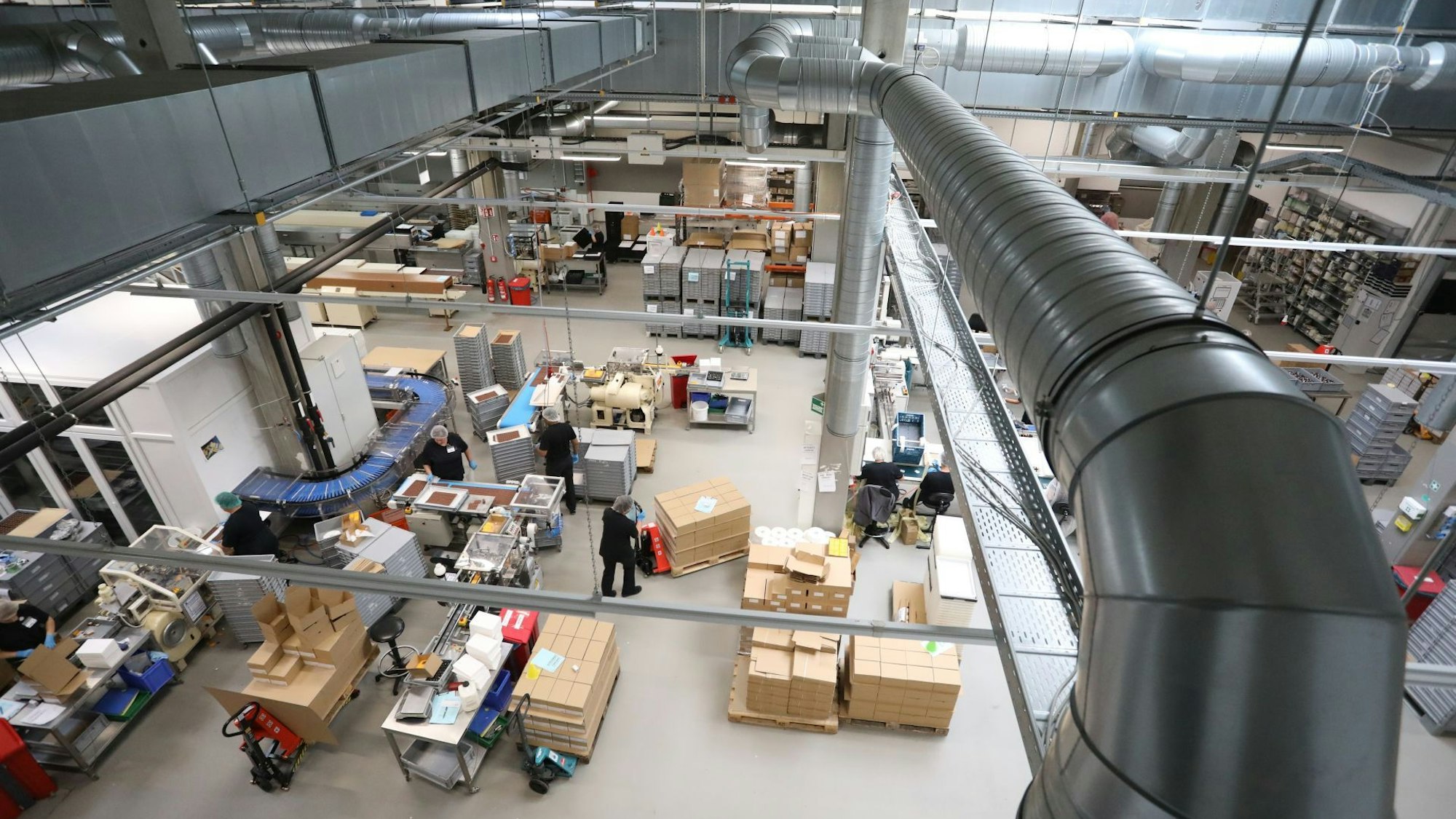 Blick von oben in die Produktionshalle der Confiserie mit diversen Apparate, Fließbändern, Kartons, Mitarbeitern und Lüftungsrohren.