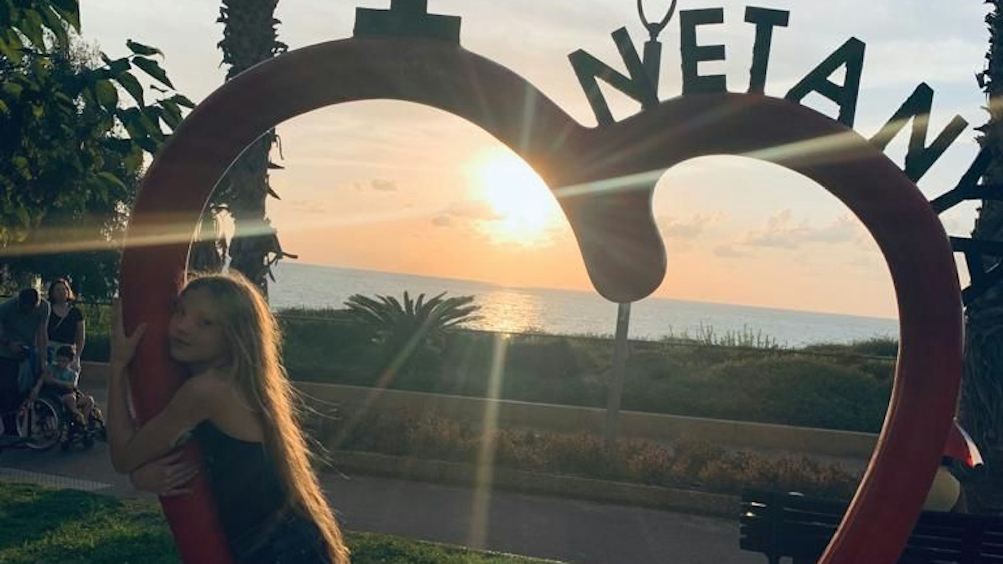 Ein Mädchen steht in einer Herzskulptur, hinter ihr geht die Sonne im Meer unter.