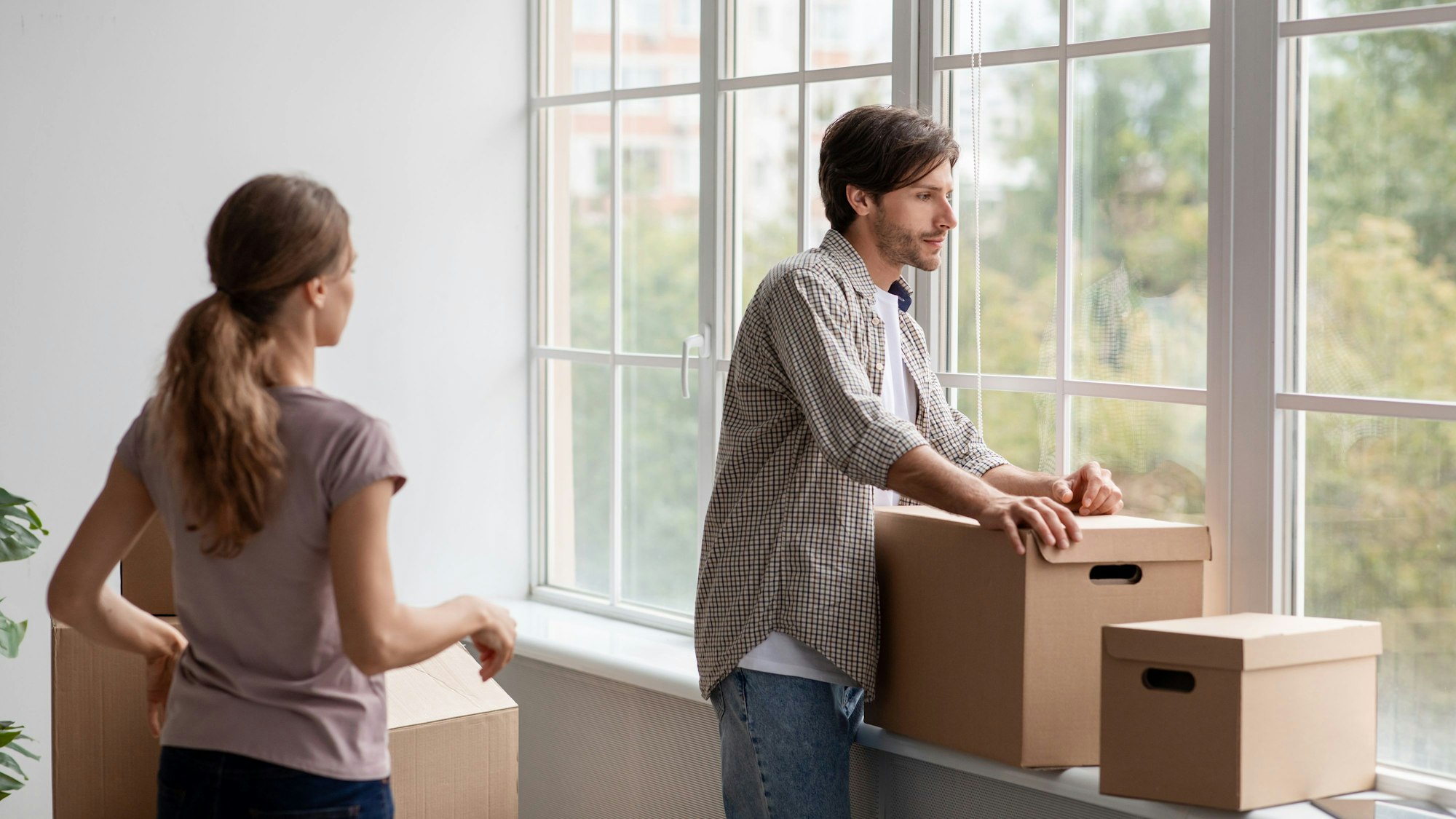 ein Mann steht mit gepacktem Umzugskarton am Fenster und schaut unglücklich hinaus, eine Frau hinter ihm versucht, mit ihm zu reden.