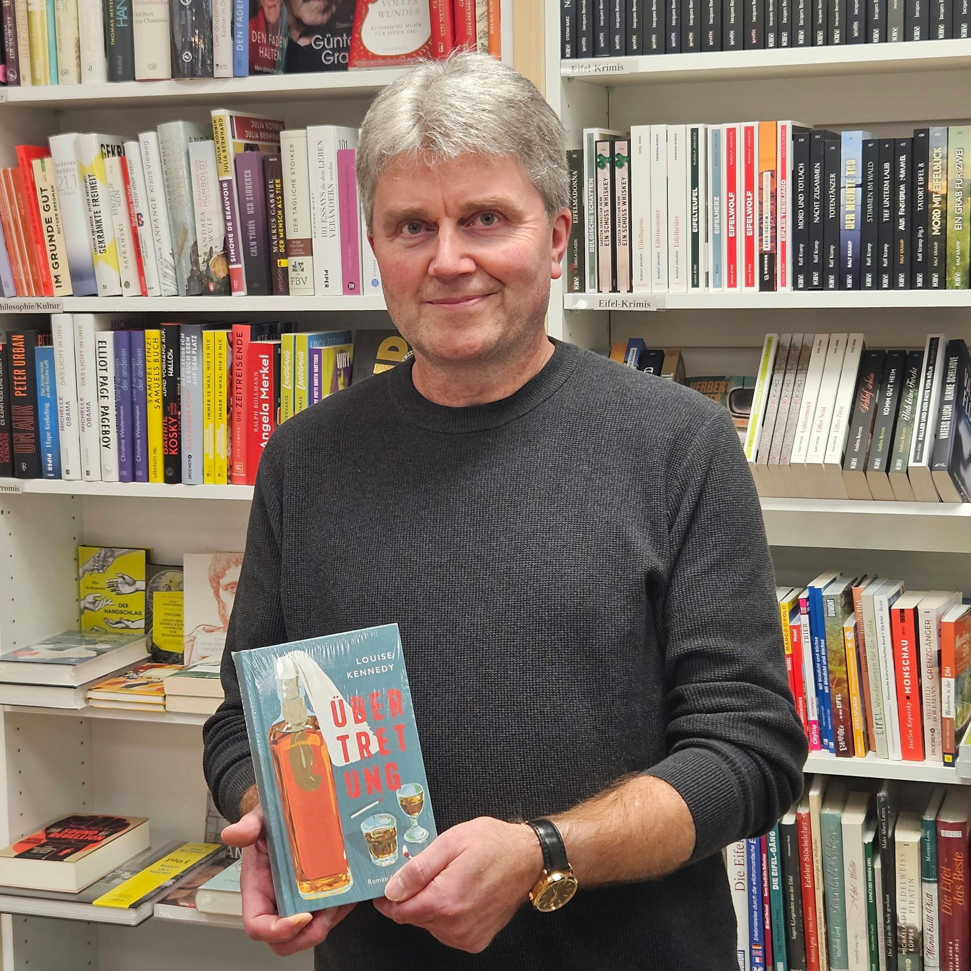 Thomas Pavlik Kall Buchtipp steht mit einem Buch von Louise Kennedy vor einem Bücherregal.